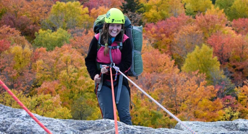 A person wearing safety gear is attached by ropes as they lean over the edge of a rock cliff, high above fall foliage below, and smile for the photo. 